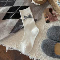 White ribbed crew sock with a gray gingham bow and broad straight cuff, shown flat beside styling props