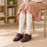 AI outfit illustration of Cream cable long socks with plain straight cuffs, an apricot skirt and burgundy Oxford shoes beside a wooden bookcase.