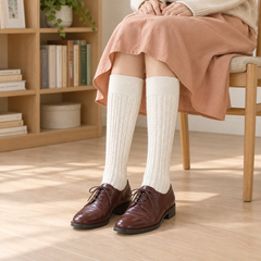 AI outfit illustration of Cream cable long socks with plain straight cuffs, an apricot skirt and burgundy Oxford shoes beside a wooden bookcase.