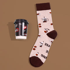 Cream coffee-themed crew sock with brown trim, coffee cups, beans and brewing tools, beside cup-shaped sock packaging