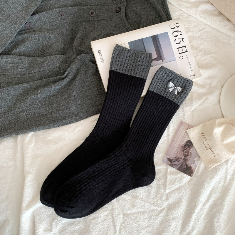 Black ribbed crew socks with broad gray cuffs and a small white embroidered bow, laid flat beside styling props