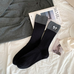 Black ribbed crew socks with broad gray cuffs and a small white embroidered bow, laid flat beside styling props