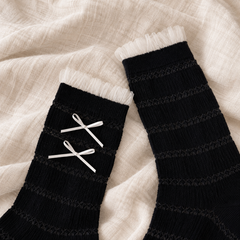 AI product illustration of Black striped-knit sock cuffs with narrow tulle edges and two thin off-white bows on the observed face, resting on oat linen.