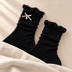 AI product illustration of two Black ruffle-cuff sock shafts on oatmeal paper, one showing its small bow and single pearl-style drop.
