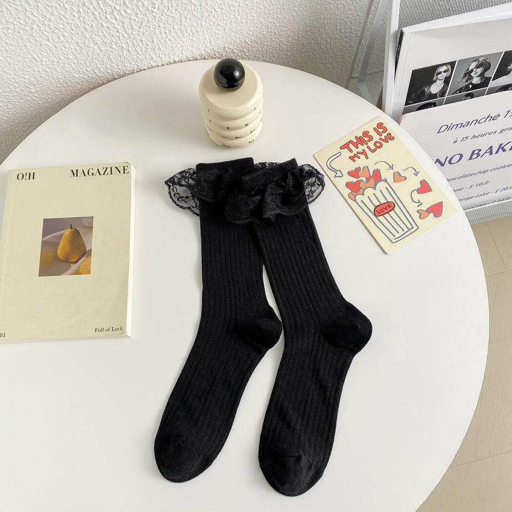 Untouched original reference of the Black ribbed lace-flounce sock flat pair, shaped heels and rounded toes; surrounding cards and objects are photo props.